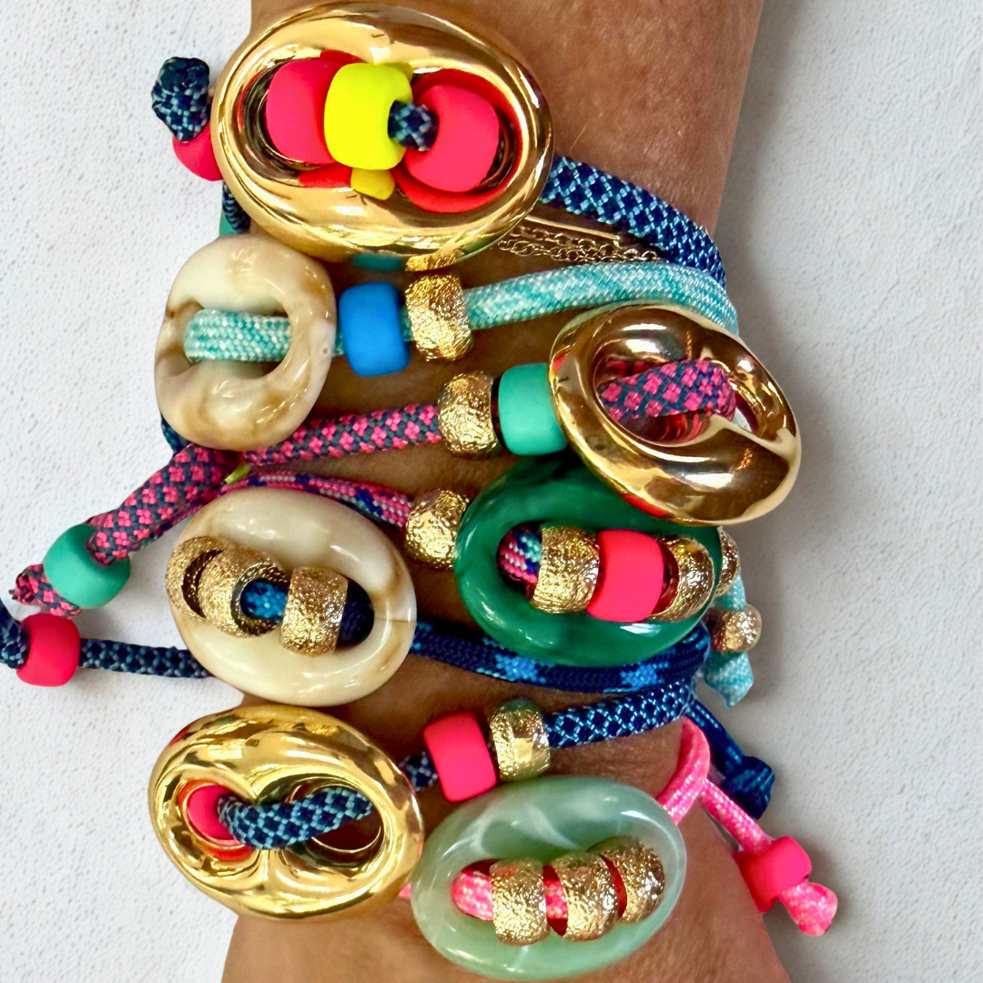 A close-up of a person's wrist wearing a stack of colorful beaded bracelets with gold-tone circular charms and braided cords in blue, pink, green, and cream.